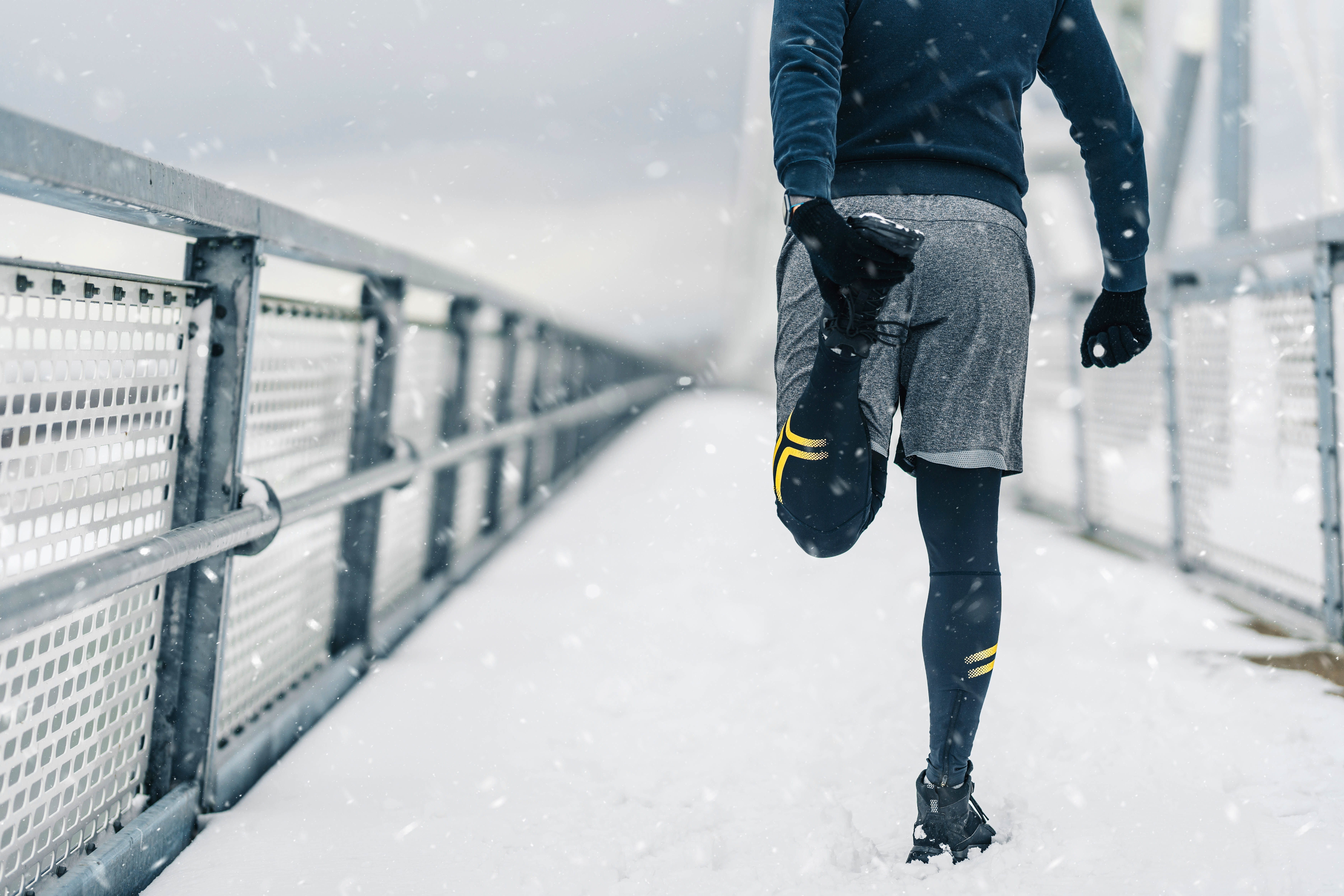 Back of a man stretching his legs and calves before heading out on a winter run across a bridge, snow is falling